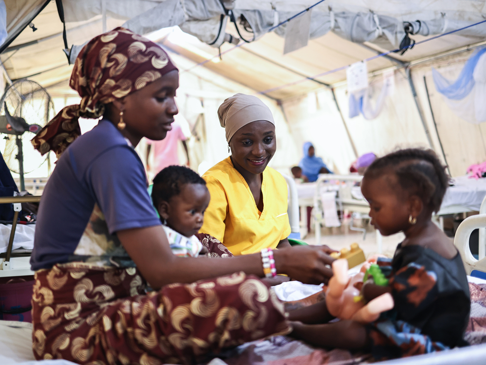 Kethurah John, conseillère et formatrice pour MSF, observe Adama jouer avec sa fille, Fatima, au centre MSF Nilefa Kiji de Maiduguri, dans l'État de Borno, au nord-est du Nigéria. Fatima est hospitalisée et reçoit un traitement contre la malnutrition. Nigeria, novembre 2025 ©Abba Adamu Musa/MSF
