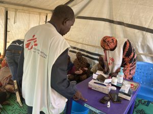 Un superviseur médical et une infirmière de MSF examinent ensemble les patients et remplissent les registres de suivi à la clinique mobile installée sur le site de transit d'Adémour, dans la province de Sila, au Tchad. À l'arrière-plan, des mères et leurs enfants attendent leur consultation médicale ou nutritionnelle.