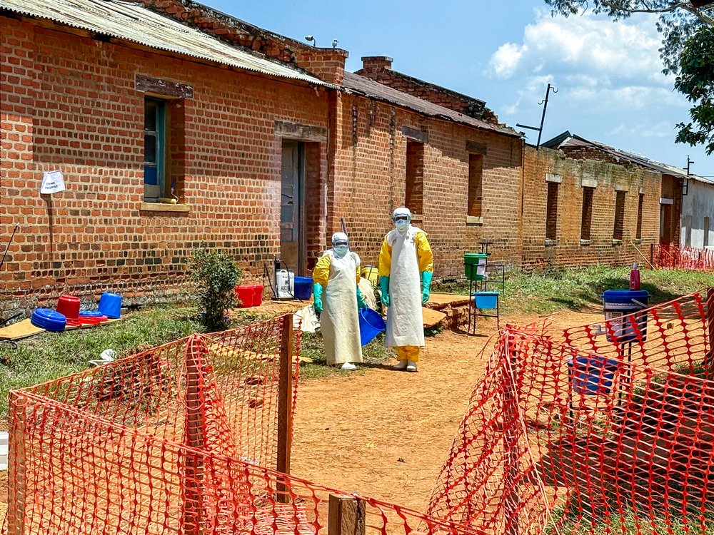 Deux agents de santé vêtus d'équipements de protection individuelle (EPI) se tiennent devant les unités de soins accueillant des patients atteints d'Ebola au centre de traitement Ebola (CTE) de MSF, situé à l'hôpital général de référence de Bambu. Ituri, Septembre 2026 ©Kristen Poels/MSF