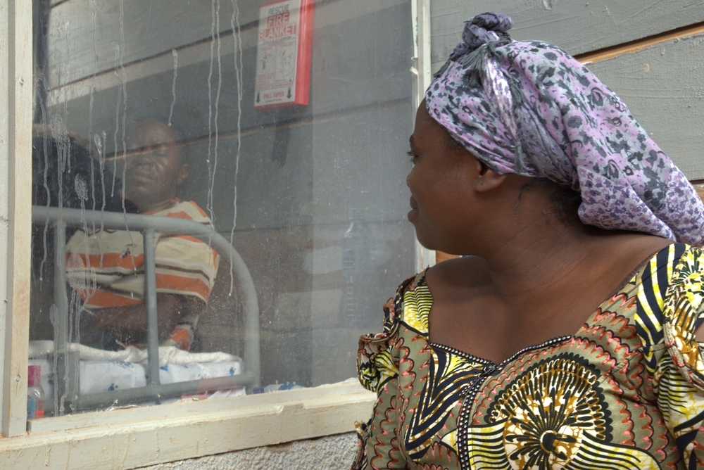 Comme elle le fait chaque jour depuis 13 jours — soit depuis l’hospitalisation de son mari, Victoire —, Kasoki a l’intention de rester à ses côtés de 6 h 30 à 18 h 30. Kasoki observe son mari de l’autre côté de la vitre et lui apporte son soutien, alors que son état de santé montre des signes d’amélioration. RDC, Butembo, Octobre 2026 ©Elio Parfait/MSF