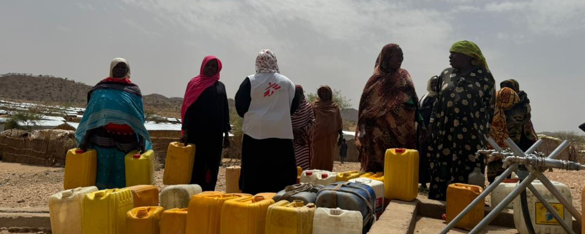 Sudanese refugee women wait at a water collection point in Metché refugee camp, eastern Chad. Rows of yellow jerrycans are placed in front of the distribution facilities, reflecting the daily importance of water access for families living in the camp.